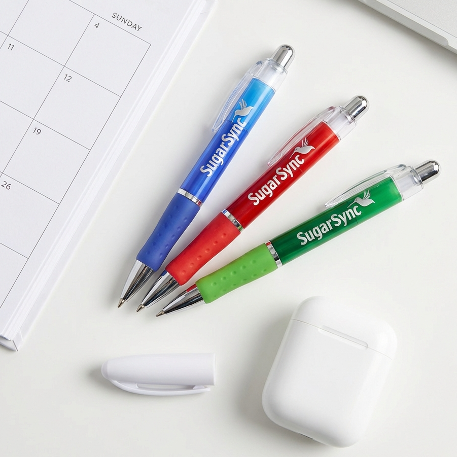 Three Translucent Xpress Odyssey pens (blue, red, green) by PensXpress rest on a white surface near a calendar, pen cap, and wireless earbud case.