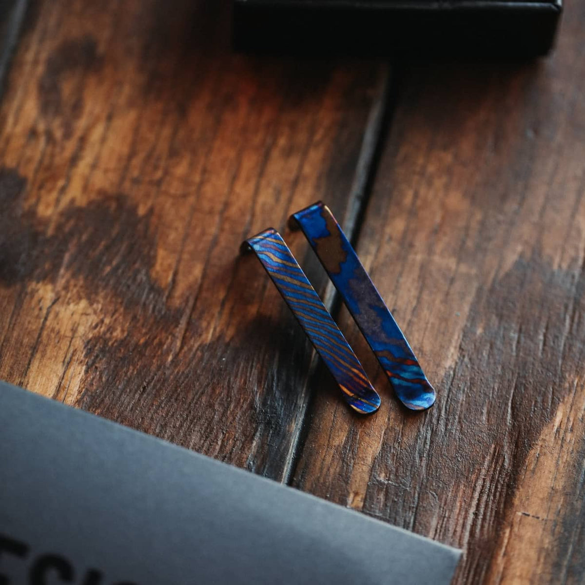 Two titanium Damascus pen clips with blue and bronze patterns on a wooden surface by Big Idea Design LLC